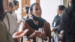 Close up of person in brown jacket and black scarf, talking with a group of colleagues